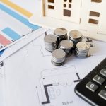 Drewniany model domu, stosy monet, kalkulator oraz wykresy finansowe leżące na planach architektonicznych, obrazujące zarządzanie funduszami inwestycyjnymi w sektorze nieruchomości i budownictwa.