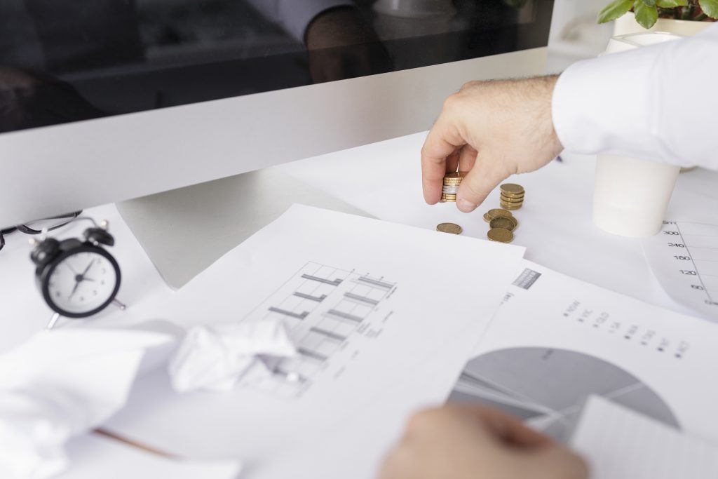 Mężczyzna przy biurku analizuje dokumenty z wykresami i układa monety, co ilustruje proces analityczny i podejmowanie decyzji, czyli aktywne zarządzanie funduszami inwestycyjnymi.